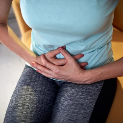 "Person clutching their stomach in discomfort, wearing a light blue shirt and gray leggings, sitting on a chair—illustrating abdominal pain or digestive issues."